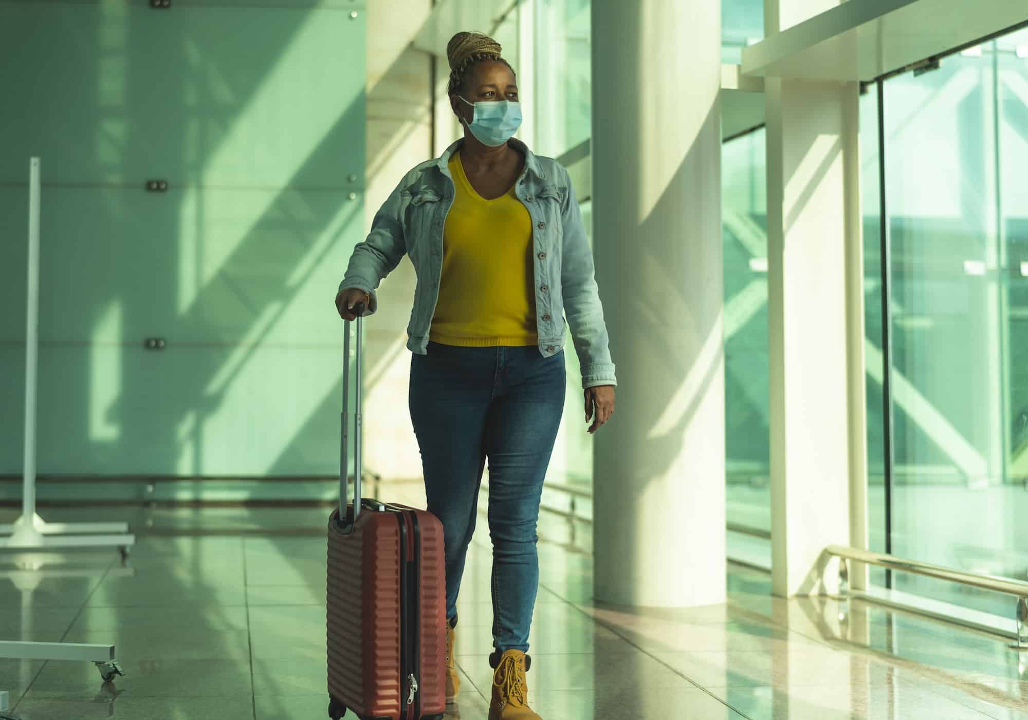 African senior woman walking in airport terminal with luggage wearing face mask